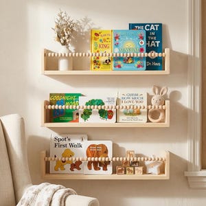 May include: Three light-colored wooden wall shelves featuring a front safety rail made of natural wooden beads. The shelves hold various children's books, a white vase with dried flowers, a crocheted rabbit rattle, and small wooden blocks.