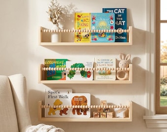 May include: Three light-colored wooden wall shelves featuring a front safety rail made of natural wooden beads. The shelves hold various children's books, a white vase with dried flowers, a crocheted rabbit rattle, and small wooden blocks.