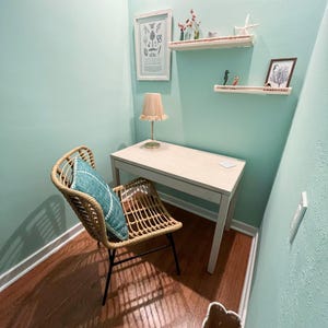 May include: A small home office space with a light wood desk, a woven chair with a blue pillow, and a beige lamp. The walls are painted a light teal color, and there are decorative shelves with small items and framed art.