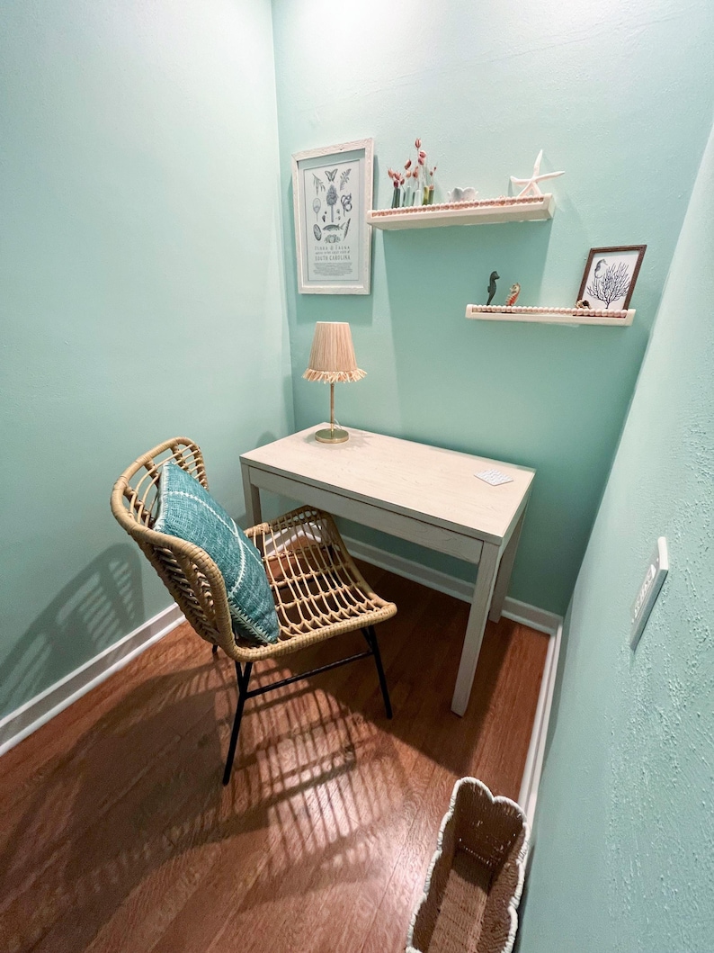 May include: A small home office space with a light wood desk, a woven chair with a blue pillow, and a beige lamp. The walls are painted a light teal color, and there are decorative shelves with small items and framed art.