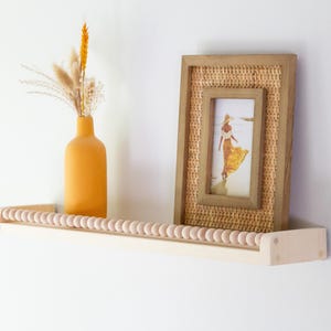 May include: A wooden shelf with a row of wooden beads along the front edge. An orange vase with dried flowers and a framed picture of a woman in a yellow dress are displayed on the shelf. The frame has a woven texture.