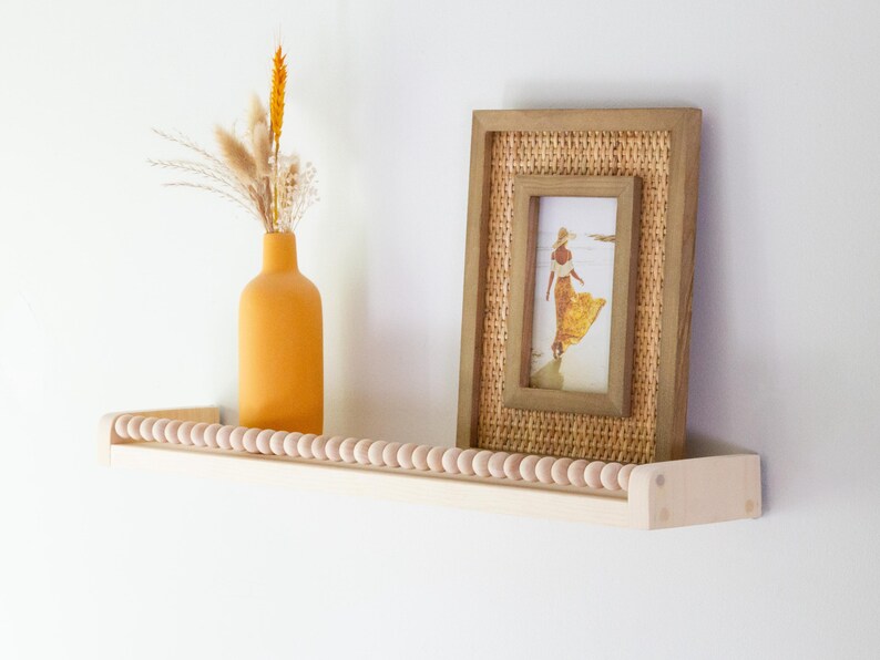 May include: A wooden shelf with a row of wooden beads along the front edge. An orange vase with dried flowers and a framed picture of a woman in a yellow dress are displayed on the shelf. The frame has a woven texture.