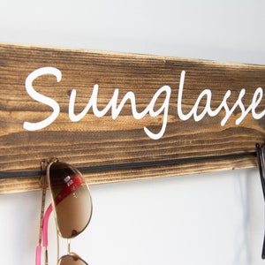 May include: Wooden wall-mounted sunglasses holder with the word "Sunglasses" written in white cursive lettering. Two pairs of sunglasses are hanging on the holder.