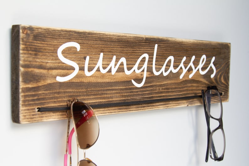 May include: Wooden wall-mounted sunglasses holder with the word "Sunglasses" written in white cursive lettering. Two pairs of sunglasses are hanging on the holder.