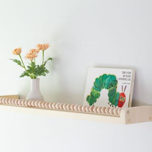 May include: A wooden shelf with a row of wooden beads along the front edge. A vase with peach-colored flowers and a copy of "The Very Hungry Caterpillar" book are on the shelf. The shelf is mounted on a white wall.