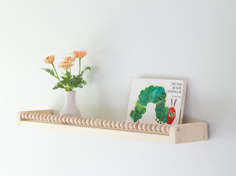 May include: A light wood shelf with a row of wooden beads along the front edge. A vase with peach-colored flowers and a copy of "The Very Hungry Caterpillar" book are on the shelf.