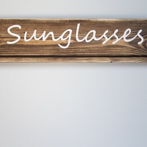May include: A wooden sign with the word "Sunglasses" painted in white on a brown background. The sign has a small shelf underneath.