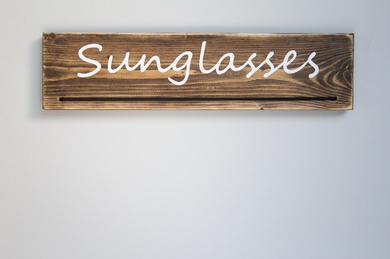 May include: A wooden sign with the word "Sunglasses" painted in white on a brown background. The sign has a small shelf underneath.