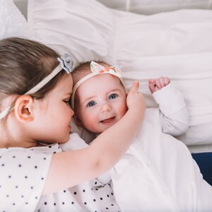 Pu&ograve; includere: Una bambina con una coda di cavallo e una camicia a pois tiene in braccio una bambina che indossa un body bianco e una fascia per capelli floreale. La bambina sorride e guarda la telecamera.