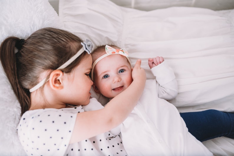 Pu&ograve; includere: Una bambina con una coda di cavallo e una camicia a pois tiene in braccio una bambina che indossa un body bianco e una fascia per capelli floreale. La bambina sorride e guarda la telecamera.