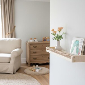 May include: A light wood floating shelf with a decorative wooden dowel edge, holding a white vase with orange flowers and a children's book. The shelf is mounted on a white wall in a nursery setting.