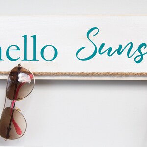 Puede incluir: Un letrero de madera blanco con el texto "hello sunshine" en letras azul turquesa. Una cuerda marr&oacute;n est&aacute; atada al letrero con dos pares de gafas colgando.
