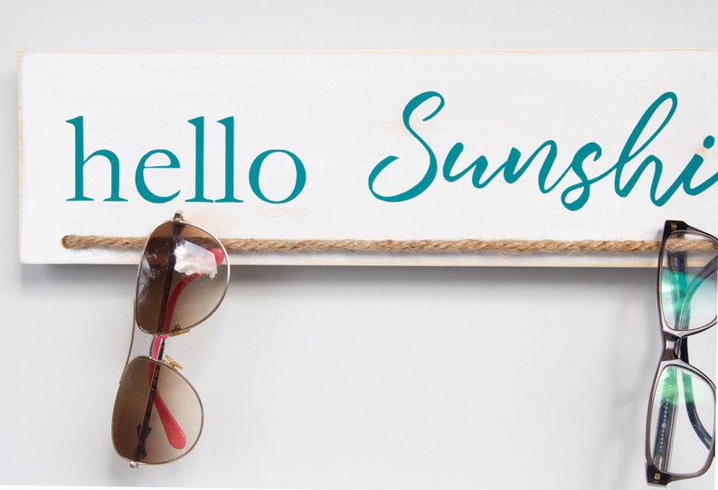 Puede incluir: Un letrero de madera blanco con el texto "hello sunshine" en letras azul turquesa. Una cuerda marr&oacute;n est&aacute; atada al letrero con dos pares de gafas colgando.
