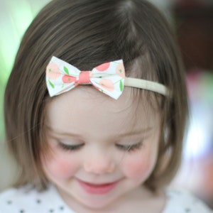 Pu&ograve; includere: Una bambina con capelli castano corti indossa una fascia bianca con un fiocco floreale rosa e bianco. Sta guardando in basso e sorridendo.