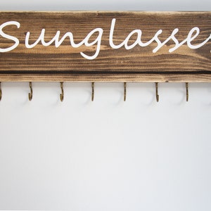 May include: A wooden wall-mounted sunglasses holder with the word "Sunglasses" painted in white. The holder has eight hooks for hanging sunglasses.
