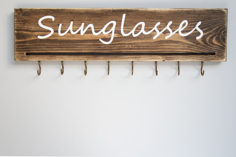 May include: A wooden wall-mounted sunglasses holder with the word "Sunglasses" painted in white. The holder has eight hooks for hanging sunglasses.