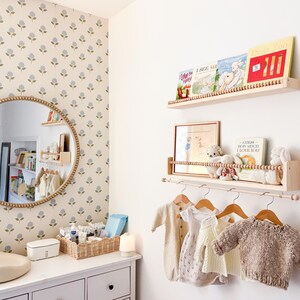 May include: A nursery room with a white dresser, a round mirror with a beaded frame, and a floral wallpaper. Wooden shelves display books and stuffed animals. Baby clothes hang on wooden hangers.