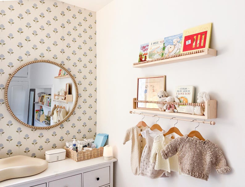 May include: A nursery room with a white dresser, a round mirror with a beaded frame, and a floral wallpaper. Wooden shelves display books and stuffed animals. Baby clothes hang on wooden hangers.