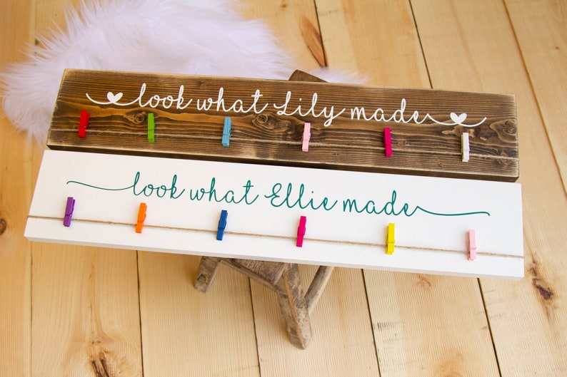 Op de afbeelding: Twee houten borden met de tekst "look what [name] made" en kleurrijke wasknijpers die aan een touwtje hangen. Het bovenste bord is bruin en het onderste bord is wit.