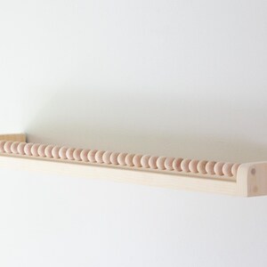 May include: A light-colored wooden shelf with a row of small, round wooden beads along the front edge. The shelf is mounted on a white wall, creating a minimalist and clean aesthetic. The shelf is designed for displaying small items.