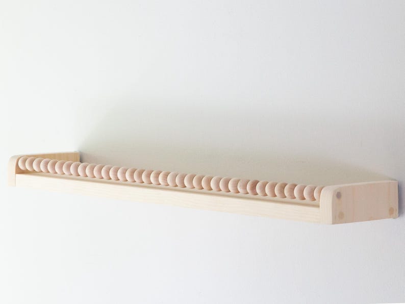 May include: A light-colored wooden shelf with a row of small, round wooden beads along the front edge. The shelf is mounted on a white wall, creating a minimalist and clean aesthetic. The shelf is designed for displaying small items.