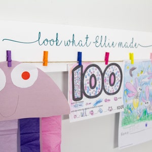 Op de afbeelding: Een wit houten bord met de tekst "look what Ellie made" in turquoise cursief. Het bord heeft een touwtje met kleurrijke wasknijpers die kunstwerken vasthouden, waaronder een paarse kwal, een tekening van het getal 100 en een tekening van vuurvliegjes.