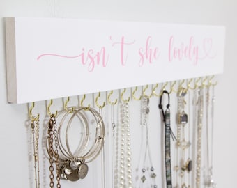Puede incluir: Un organizador de joyas de pared rectangular de madera blanca con el texto en cursiva rosa "isn't she lovely" y un corazón. El tablero incluye una fila de ganchos de metal dorado para colgar varios collares, pulseras y cadenas.