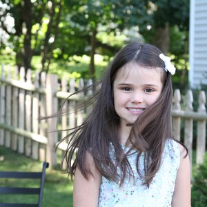 Pu&ograve; includere: Una bambina con lunghi capelli castani sorride alla telecamera. Indossa un abito blu chiaro con paillettes e un fiore bianco nei capelli.