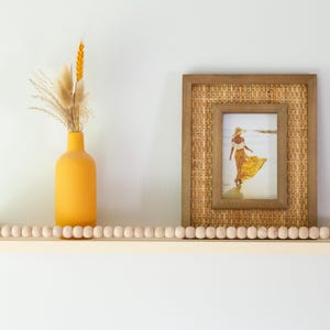 May include: A wooden shelf with a beaded edge, holding a yellow vase with dried flowers and a framed picture. The picture has a woven border and shows a person in a yellow dress on a beach.