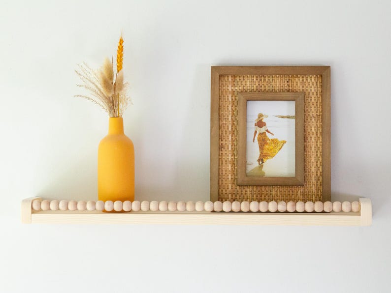 May include: A wooden shelf with a beaded edge, holding a yellow vase with dried flowers and a framed picture. The picture has a woven border and shows a person in a yellow dress on a beach.