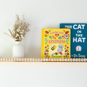 May include: A wooden shelf with a beaded edge holds two children's books, "Kindness" and "The Cat in the Hat." A white vase with dried flowers sits to the left. The shelf is mounted on a white wall.