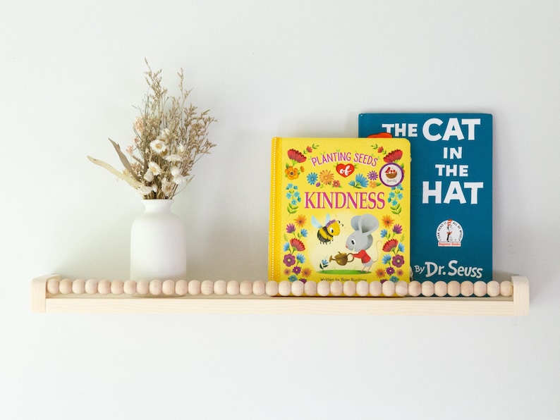 May include: A wooden shelf with a beaded edge holds two children's books, "Kindness" and "The Cat in the Hat." A white vase with dried flowers sits to the left. The shelf is mounted on a white wall.