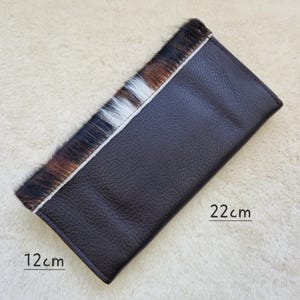 May include: A dark brown leather wallet with a section of cowhide hair in shades of brown, black, and white. The wallet measures 4.7 inches wide and 8.7 inches long.