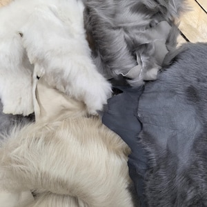 May include: A collection of multiple pieces of faux fur fabric in various shades including white, light beige, medium grey, and dark charcoal grey. Each piece displays a soft, long-pile texture with visible smooth backing material on the edges.