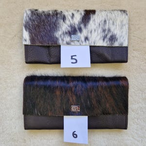 May include: Two leather wallets with cowhide accents. The top wallet has a white and brown pattern, while the bottom wallet has a dark brown and black pattern. Both wallets have a dark brown leather base and a silver clasp. The number 5 and 6 are on white tags.