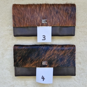 May include: Two leather wallets with a cowhide flap. One wallet has a brown and tan cowhide flap, and the other has a black and brown cowhide flap. Each wallet has a dark brown leather base and a silver emblem. The number "3" and "4" are on white tags.