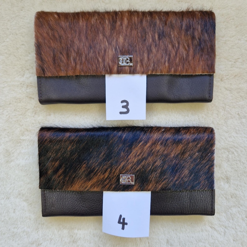 May include: Two leather wallets with a cowhide flap. One wallet has a brown and tan cowhide flap, and the other has a black and brown cowhide flap. Each wallet has a dark brown leather base and a silver emblem. The number "3" and "4" are on white tags.