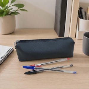 May include: A dark navy blue rectangular pencil case with a zipper closure. The case is positioned horizontally on a surface with three ballpoint pens lying in front of it, featuring red, blue, and black caps and ink cartridges respectively.