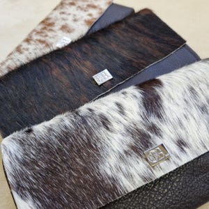 May include: Close-up of three leather wallets with cowhide accents. The wallets feature a variety of colors, including brown, black, and white. Each wallet has a silver clasp. The wallets are rectangular and have a flap closure.