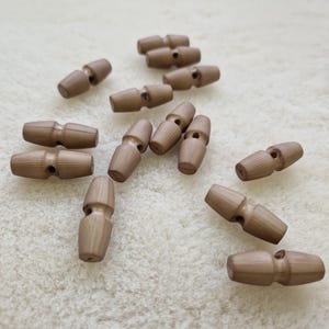 May include: A selection of light brown wooden toggle buttons, each with a barrel shape and vertical grooves. The buttons are scattered on a white, textured surface. Suitable for coats and jackets.