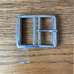 May include: A square, silver metal buckle with a rectangular frame and three vertical bars. The buckle is set against a wooden background with visible grain.