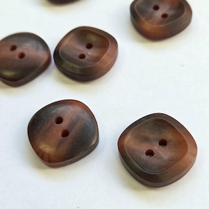 May include: Close-up of several brown, square-shaped buttons with rounded corners. Each button has two holes and a marbled appearance, with shades of brown and a darker rim. The buttons are scattered on a white surface.