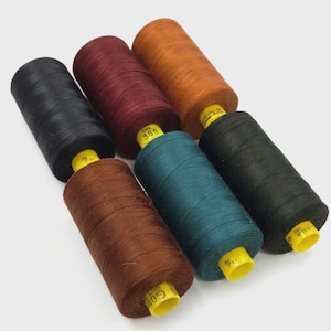 May include: Six cylindrical spools of sewing thread arranged in two rows. The colors include black, deep burgundy, burnt orange, brown, teal, and a second black spool. Each spool features a bright yellow plastic core with printed text.