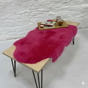 May include: A vibrant pink sheepskin rug on a light wood table with black hairpin legs. A wooden tray holds a mug, a muffin, and a book. The rug adds a bold colour statement to the room.