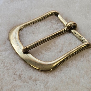 May include: A gold-colored metal belt buckle with a rounded rectangular shape. The buckle has a single bar and a pin for securing a belt. The metal has a slightly aged appearance.