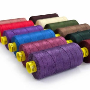 May include: A collection of twelve cylindrical spools of sewing thread arranged in a grid. The spools feature vibrant colors including purple, blue, red, pink, cream, olive green, brown, and dark green, each with a bright yellow plastic core.