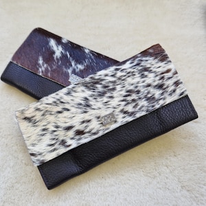 May include: Two leather wallets with cowhide accents. One wallet features a brown and white spotted pattern, while the other has a dark brown and white pattern. Both wallets have a black leather base and a silver-colored clasp.