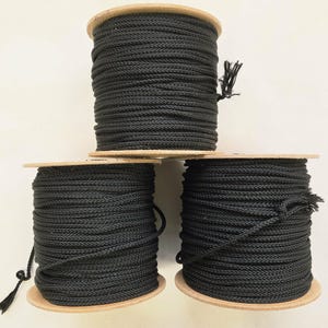 May include: Three spools of black rope are stacked on top of each other. The rope is tightly wound around wooden spools. The rope has a braided texture. The spools are on a light background.