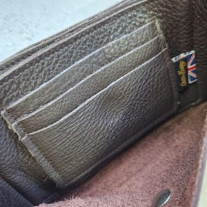 May include: Open brown leather wallet with multiple card slots and a zippered compartment. The wallet has a textured surface and a small tag with the text "Owen Barry" and a Union Jack flag.