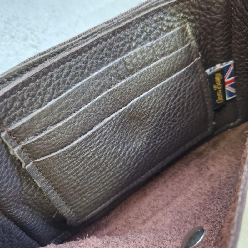 May include: Open brown leather wallet with multiple card slots and a zippered compartment. The wallet has a textured surface and a small tag with the text "Owen Barry" and a Union Jack flag.
